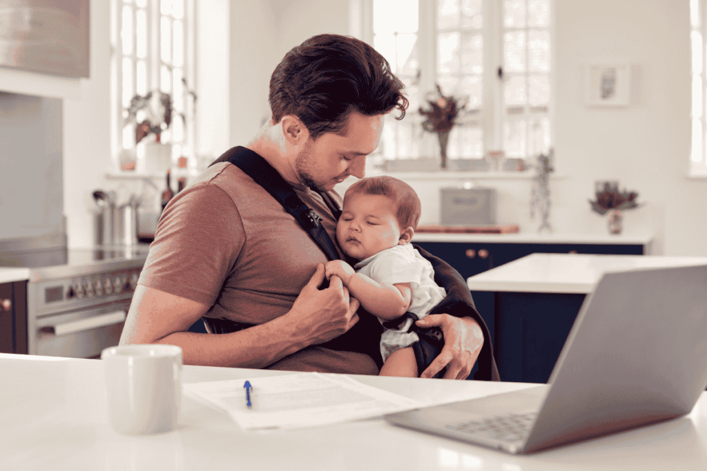 Vater arbeitet am Laptop, während sein Baby in der Trage auf seiner Brust schläft – Symbolbild für die Einschlafbegleitung durch Papa oder Oma